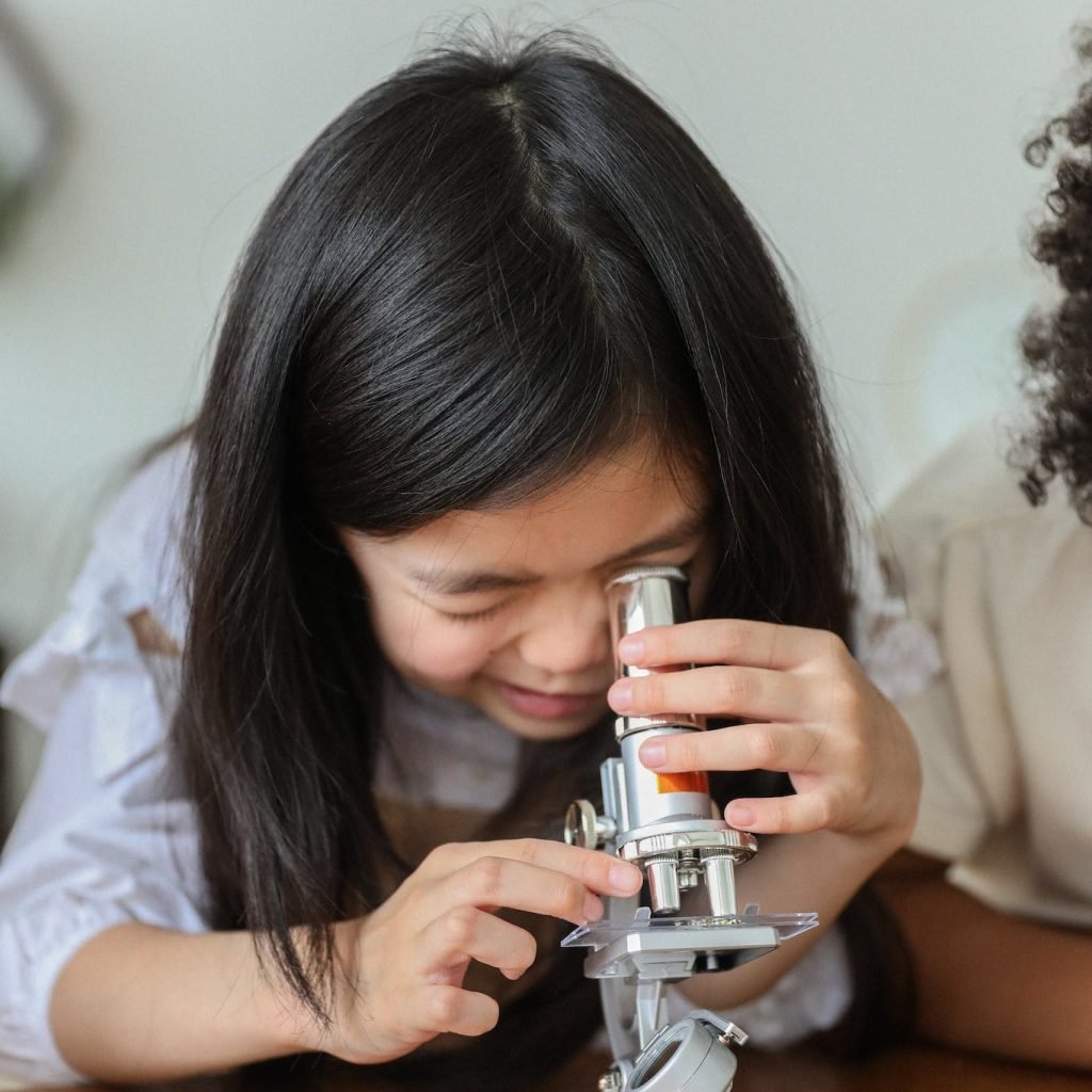 Cute little Asian girl with long dark hair making experiment with microscope while sitting at wooden table in classroom